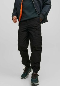 Pantaloni cargo neri con polsini elasticizzati, dotati di tasche laterali e un fit rilassato. Indossati con una giacca scura e sneakers in materiali misti.