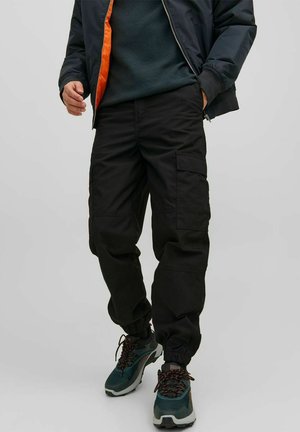 Pantaloni cargo neri con polsini elasticizzati, dotati di tasche laterali e un fit rilassato. Indossati con una giacca scura e sneakers in materiali misti.