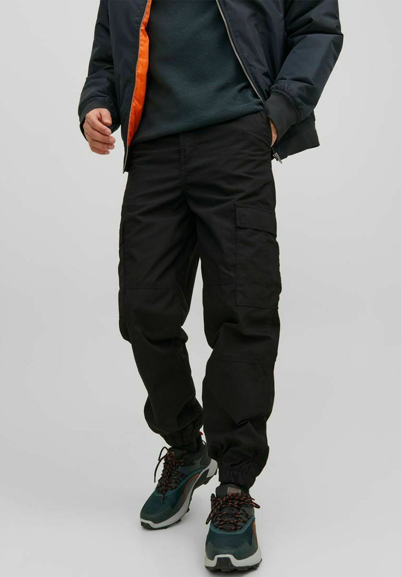 Pantaloni cargo neri con polsini elasticizzati, dotati di tasche laterali e un fit rilassato. Indossati con una giacca scura e sneakers in materiali misti.