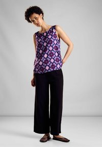 Top senza maniche viola con motivo geometrico in rosa e bordeaux, abbinato a pantaloni neri a gamba larga e scarpe nere piatta.