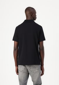 Melnas īsām piedurknēm polo krekls ar plakanu apkakli, izgatavots no gludas kokvilnas auduma, saskaņots ar pelēkiem džinsu biksēm, kas demonstrē ikdienišķu piegriezumu.