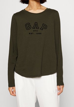 Modèle portant un haut Gap à manches longues, vert olive foncé, avec des lettres noires, et un pantalon blanc.