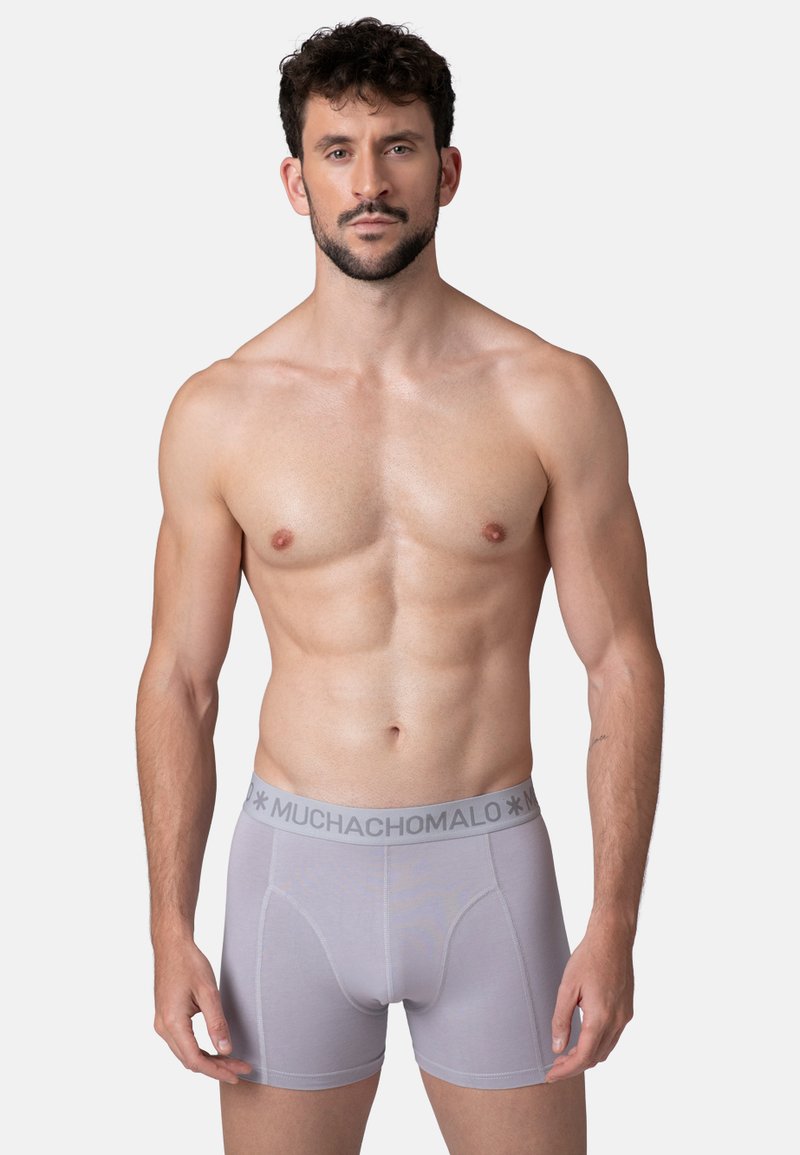 Hombre musculoso con cabello rizado oscuro, vistiendo bóxer gris claro con cinturilla "MUCHACHOMALO", de pie contra un fondo blanco liso.