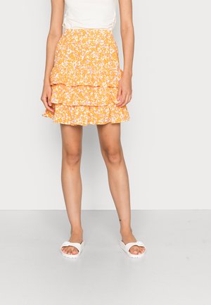 Saia floral amarela com três camadas, apresentando detalhes em branco e lilás. O material parece leve, com uma bainha encorpada. Usada com sandálias brancas.