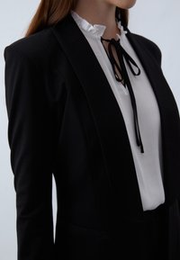 Czarny blezer o dopasowanym kroju, z klapami z wycięciem i kieszenią. Pod spodem biała bluzka z marszczonym kołnierzem i czarnymi kokardami.