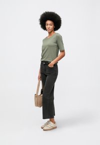 Γυναίκα με αφρο χτένισμα, φοράει πράσινη μπλούζα με κοντά μανίκια, μαύρα cropped τζιν, λευκά sneakers, κρατώντας μπεζ τσάντα, στέκεται μπροστά σε απλό φόντο.