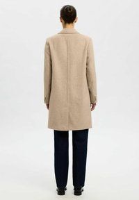 Lange beige wollen jas met een gestructureerd ontwerp, een enkelvoudige sluiting en subtiele schouderaccenten, gecombineerd met donkere spijkerbroek en zwarte schoenen.