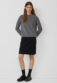 Grijs shirt met lange mouwen en een ronde hals, gecombineerd met een zwarte rok tot op de knie. Zwarte schoenen met dikke zolen en donkere sokken maken de outfit compleet.