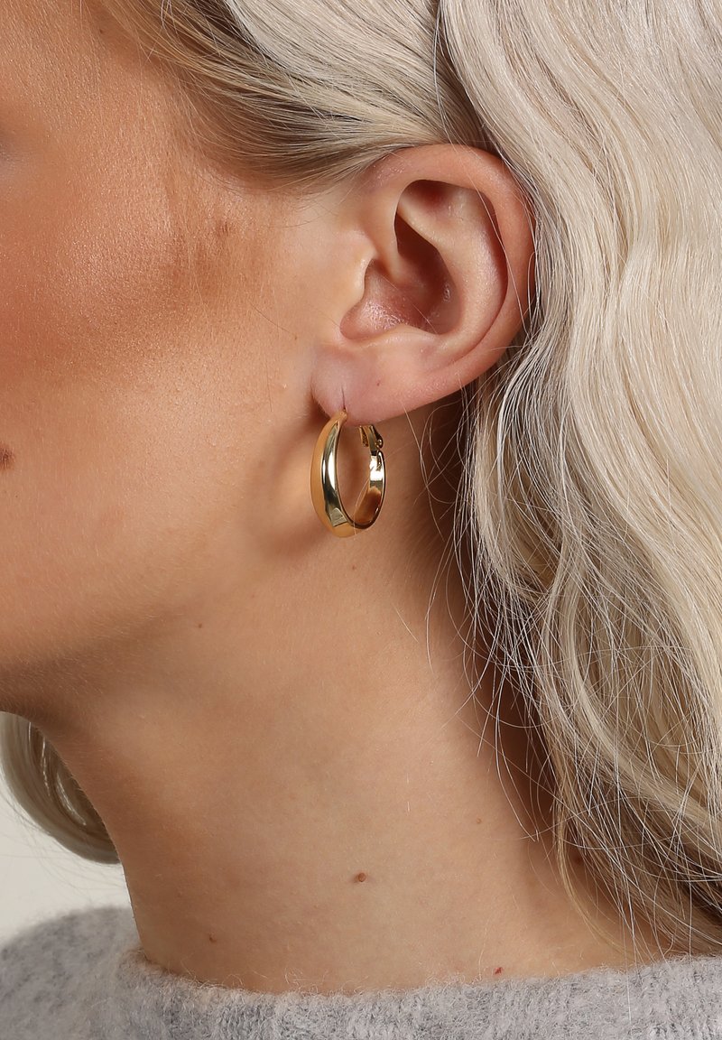 Guldörhänge med en slät, glänsande yta och en lätt kurva, visad på en modells öra, vilket framhäver dess minimalistiska design.