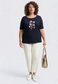 Granatowa koszulka z krótkim rękawem z kwiatową haftem na przodzie. Połączona z beżowymi spodniami i trzymająca beżową torbę. Białe trampki.