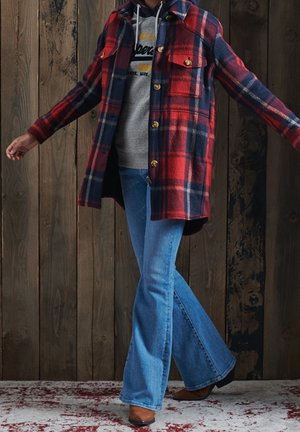 Rote und marineblaue Kariertes Flanelljacke mit zwei Brusttaschen, Knopfverschluss und kontrastierendem Innenfutter, darüber ein grauer Hoodie und blaue Schlagjeans.