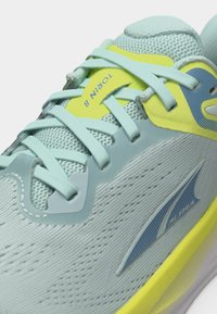 Κοντινή λήψη αθλητικού παπουτσιού σε ανοιχτό μπλε και κίτρινο χρώμα, με κορδόνια, επάνω μέρος από διχτυωτό υλικό και την επιγραφή "Torin 8" στη γλώσσα.