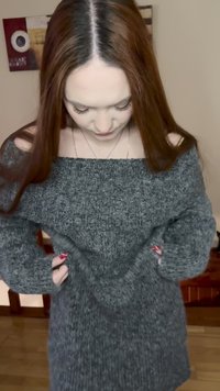 Pikkade punaste juustega naine kohandamas halli õlata kampsunit, vaadates alla, käed peitu kampsunis vöökoha taskutesse.