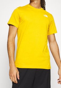 Světlé žluté bavlněné tričko s krátkým rukávem a kulatým výstřihem. Na levé straně hrudi se nachází malé bílé logo. Jednoduchý design, měkká textura.