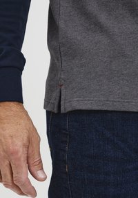 Szürke, hosszú ujjú ing, tengerészkék mandzsettákkal, enyhe oldalsó hasítékkal és piros varrással. Sötét farmerral viselve.