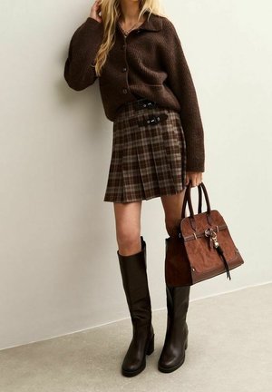 Femme portant un cardigan marron boutonné, une jupe plissée à carreaux avec des boucles, des bottes noires montantes jusqu'aux genoux, tenant un sac à main en cuir marron, debout contre un mur.