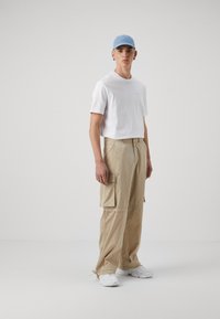 Hvit t-skjorte med korte ermer, beige cargo-bukser med sidelommer, blå caps, hvite joggesko, står mot en ensfarget bakgrunn.