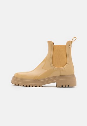 Botte Chelsea beige brillante avec des panneaux élastiques sur les côtés, languette à l'arrière et semelle crantée épaisse, vue de l'extérieur sur fond blanc.