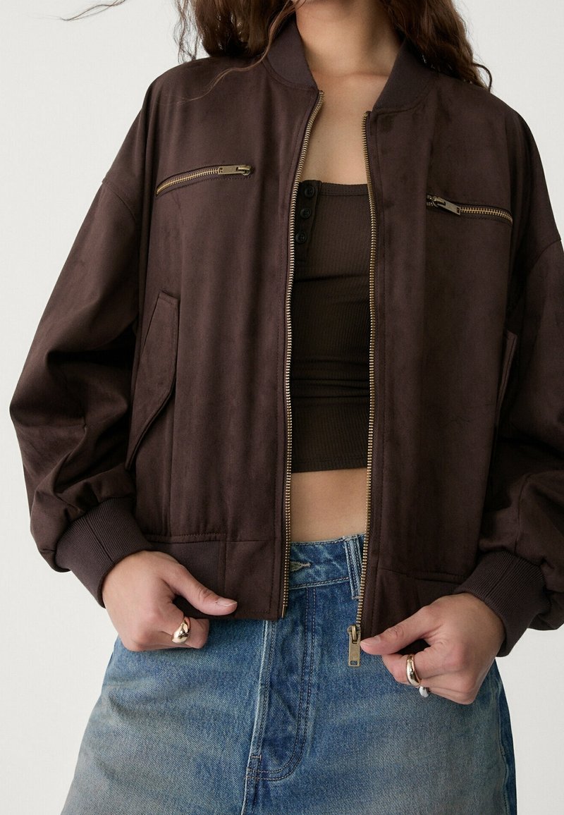 Personne portant une veste bomber marron foncé sur un top court assorti et un jean bleu en denim taille haute, les mains reposant près de la fermeture éclair.