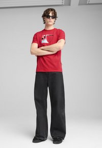 Camiseta de algodón roja con diseño gráfico que presenta formas en azul, verde y rosa. Combinada con unos pantalones anchos negros y zapatos negros.