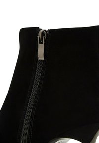 Botín de ante negro con cremallera lateral y tirador plateado, tacón alto y textura suave. Diseño minimalista y silueta elegante.