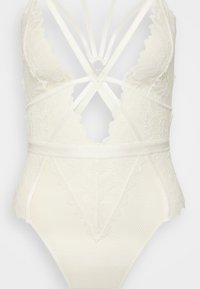 Fehér csipke body mélyen kivágott nyakkivágással, keresztezett pántokkal és texturált alsó résszel virágmintás díszítéssel. Arany színű kör alakú fém részlet.