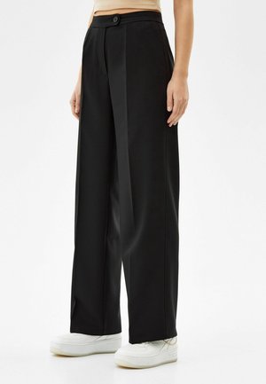 Personne portant un pantalon noir taille haute à jambes larges avec fermeture par bouton et zip, associé à des baskets blanches à plateforme sur fond blanc.