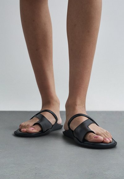 Paljain jaloin mustat, avoimet sandaalit, joissa leveät remmit, betonilattialla vaaleanharmaata taustaa vasten.