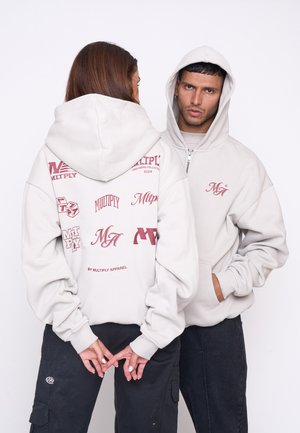 Lys grå oversized hoodie med grafiske tryk i burgundy. Designet inkluderer lynlås foran, stor hætte og ribbede manchetter.
