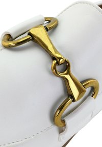 Witte leren schoen met een grote gouden hardwareaccent met lussen en een driehoekige vorm, die een gladde, textuur heeft.