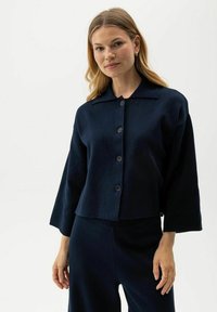 Maglione corto button-up blu navy con colletto, caratterizzato da maniche larghe e una consistenza morbida, indossato sopra pantaloni abbinati.