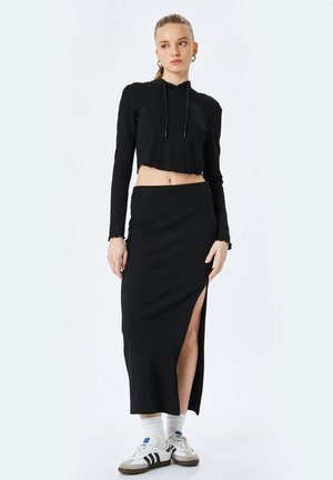 Sudadera corta negra con cordones, combinada con una falda midi negra de canalé con una abertura lateral. La modelo lleva calcetines deportivos blancos y zapatillas.