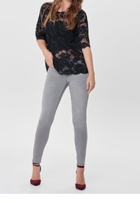 Schwarze Spitzenbluse mit Dreiviertelärmeln, kombiniert mit grauen Skinny-Jeans und bordeauxroten High-Heels. Glatte Textur, taillierter Schnitt.