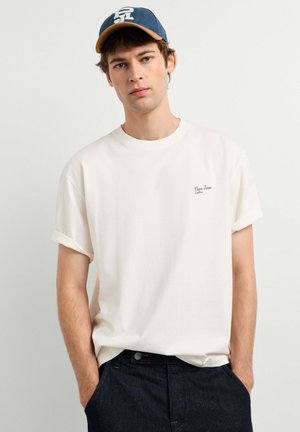 Giovane uomo che indossa una T-shirt bianca con le maniche arrotolate con il testo "Pepe Jeans London" e un berretto da baseball blu, mani nelle tasche di jeans scuri contro un muro bianco.