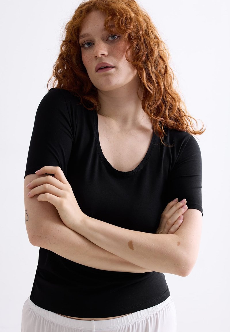 Zwart T-shirt met korte mouwen, ronde halslijn, zachte textuur en aansluitend ontwerp. Gecombineerd met witte broeken, met zichtbare huidskleur en sproeten.