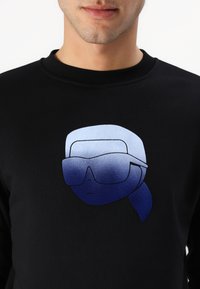 Black sweatshirt features a graphic of a stylized face with sunglasses, in blue gradient tones, with a smooth texture and crew neck design.