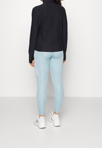 Femme debout, portant un haut noir à manches longues, un legging de sport bleu clair avec des rayures blanches, et des baskets blanches avec des chaussettes blanches.