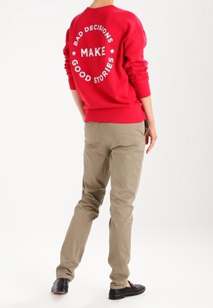 Persoon draagt een rode trui met de tekst "Slechte beslissingen zorgen voor goede verhalen" op de achterkant, beige broek en zwarte schoenen, staand tegen een witte achtergrond.