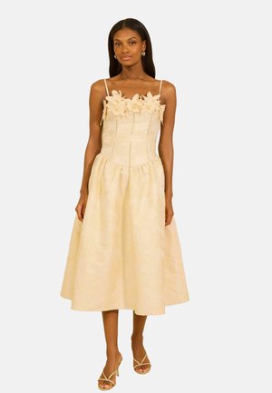 Kobieta w bladżóto-m sukience midi z kwiatową aplikacją na górze i cienkimi ramiączkami, nosząca pasujące sandały na obcasie.