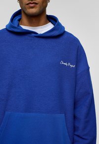 Blauwe hoodie met een gestructureerde afwerking, voorzien van een kangoeroezak en witte borduursel "Eternity Project" op de borst.