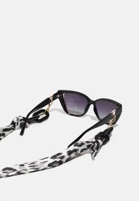 Kattøye-solbriller med svarte plastrammer, lilla gradientlinser og gull detaljer, festet til et svart og hvitt leopardmønstret bånd.