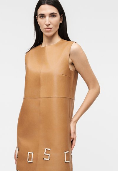 Γυναίκα που φοράει αμάνικο δερμάτινο φόρεμα σε ταμπά χρώμα με γράμματα σε στυλ σχοινιού που σχηματίζουν τη λέξη "MOSC" κοντά στο τελείωμα, στέκεται μπροστά σε απλό φωτεινό φόντο.