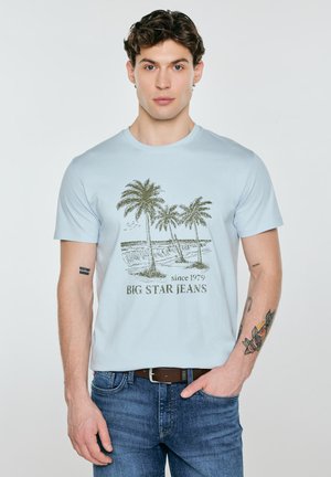 Muž nosící světle modré tričko s potiskem palmy a nápisem „Big Star Jeans“, doplněné modrými džíny a hnědým pásem.