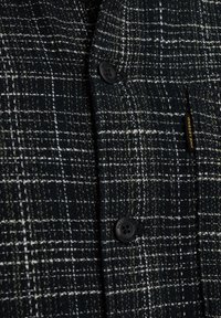 Camisa de cuadros en blanco y negro con una tela texturizada. Presenta un botón negro y una pequeña etiqueta de la marca en el bolsillo del pecho.