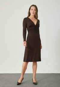 Robe marron à manches longues, décolleté en V, buste froncé et ourlet à hauteur des genoux. Texture de tissu lisse et silhouette ajustée.