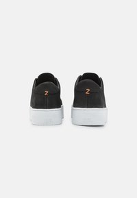 Ein Paar schwarze Wildledersneaker mit weißen dicken Sohlen, einem orangen "Z"-Logo an der Ferse und "HUB" in die Rückseite der Sohle eingeprägt.
