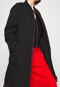 Veste bomber noire matelassée avec fermeture éclair. Comprend des poignets côtelés et un col montant, associée à un vêtement inférieur rouge. Texture lisse.