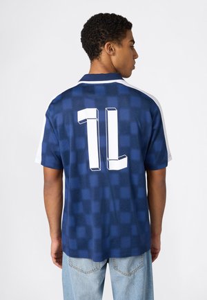 Młody mężczyzna zwrócony tyłem, ubrany w niebieską, kraciastą koszulę z krótkim rękawem, z dużym białym numerem "1L" na plecach oraz jasnoniebieskie dżinsy.