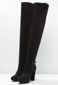 Botas de muslo negras con tacones altos, punteras redondeadas, tacones de bloque y una pequeña correa con hebilla cerca del tobillo sobre una superficie blanca.