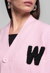 Růžový pletený svetr s texturovanou látkou, zdobený černou aplikací ve tvaru "W" a dvěma růžovými knoflíky na přední straně.